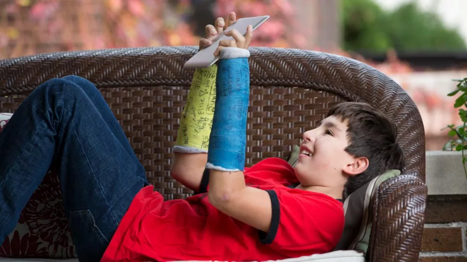 Boy with two arm casts laying on back and looking at ipad