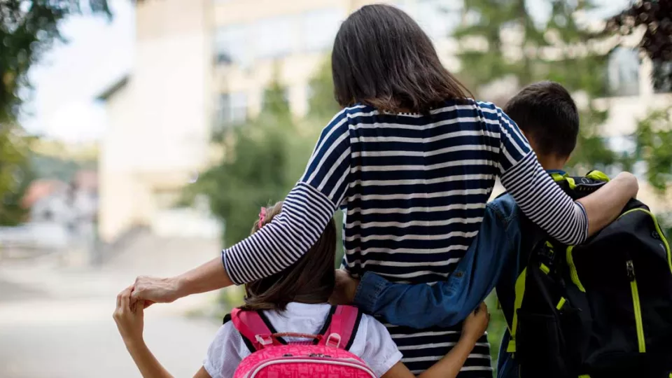 Mother with shoulder-length brown hair and striped shirt stands with her back to the camera and her arms around son and daughter