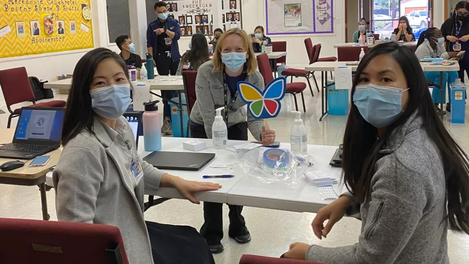 A seated medium-light skin toned woman, a standing light skin-toned woman holding a butterfly sign, and a seated medium skin-toned woman, all wearing surgical masks, smile at the camera while nurses administer vaccinations behind them