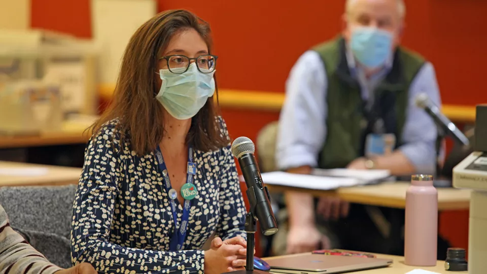 A light skin-toned woman wearing a surgical mask sits in front of a microphone. A light skin-toned man in a surgical mask behind her listens.