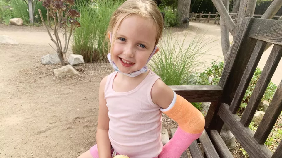 Little girl with light skin tone and left arm in a cast smiles as she sits on a wooden park bench