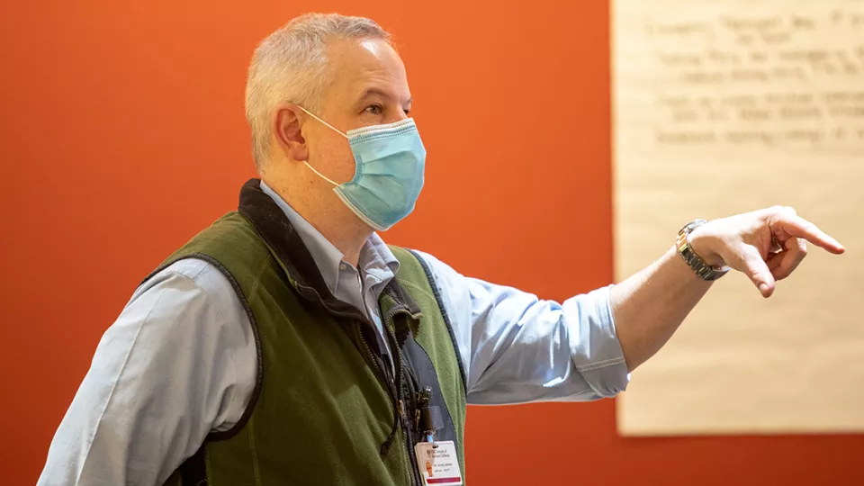 A light skin-toned man wearing a surgical mask gestures with his hand and talks.