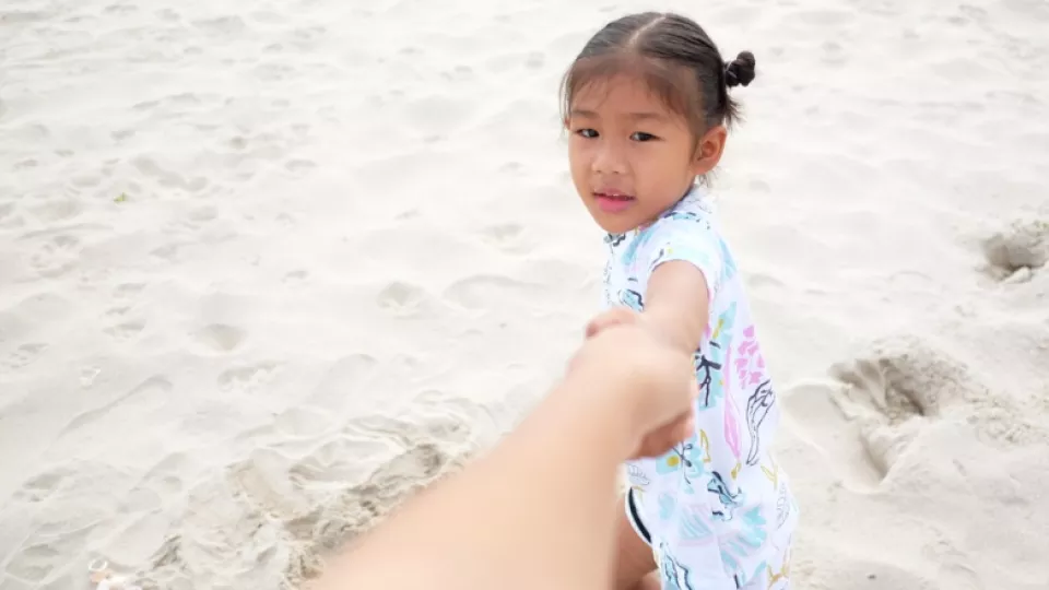 Young girl with medium skin tone and dark hair pulls her mother's arm on the beach