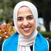 Headshot of a smiling woman with medium skin tone wearing a light colored headscarf and blue graduation stole against a blurred outdoor background