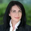 Headshot of a smiling woman with light skin tone and shoulder-length dark hair wearing a white blouse under a dark suit jacket against a blurred green outdoor background