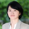 Headshot of a woman with light skin tone and short dark hair wearing a white blouse under a grey suit jacket against a blurred green outdoor background