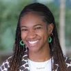 Headshot of a young woman with dark skin tone and long dark hair wearing green hoop earrings and a patterned jacket with a blurred outdoor background