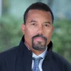 Headshot of a man with dark skin tone and dark hair wearing a blue tie and dark blue jacket against a blurred outdoor background