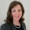 Headshot of a smiling woman with light skin tone and brown hair wearing a pearl necklace and dark suit jacket against a neutral background