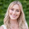 Headshot of a young woman with light skin tone and light hair wearing a white top against blurred outdoor background