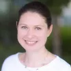 Headshot of a smiling woman with light skin tone and dark hair wearing a white top and silver heart chain necklace against a blurred outdoor background