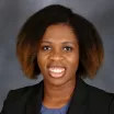 Headshot of a smiling woman with dark skin tone and dark hair wearing a blue blouse and dark suit jacket against a neutral indoor background