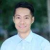 Headshot of smiling young man with medium skin tone and dark hair wearing button-down blue dress shirt against a blurred outdoor background