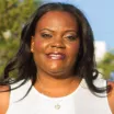 Headshot of a smiling woman with dark skin and shoulder-length dark hair wearing a white top and gold heart necklace against a blurred outdoor background