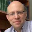 Headshot of a balding man with light skin tone and glasses wearing a open collar dress shirt with a bookcase in the background