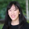 Headshot of a smiling woman with light skin and dark hair wearing a black top against a blurred outdoor background