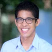 Headshot of a smiling man with medium skin tone and dark hair wearing tortoise shell glasses and an open-collar light blue dress shirt against a blurred outdoor background