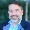 Headshot of a smiling man with light skin tone and a grey beard wearing an open-collar, light blue dress shirt under a dark blue suit jacket against a blurred outdoor background