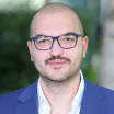 Headshot of a smiling man with light skin tone and shaved head and mustache wearing blue-framed glasses and a white, open-collar dress shirt under a blue suit jacket against a blurred outdoor background