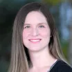 Headshot of a smiling woman with light skin tone and light brown hair wearing a black cardigan against a blurred outdoor background