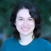 Headshot of a smiling woman with light skin tone and shoulder-length dark hair wearing a blue sweater against a blurred outdoor background