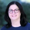 Headshot of a smiling woman with shoulder-length dark hair wearing a dark blue top and eyeglasses with a purple frame against a blurred outdoor background