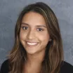 Headshot of a smiling woman with medium skin tone and straight brown hair wearing a dark top against a neutral indoor background