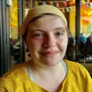 Portrait of a nonbinary adult with a closed-mouth smile and short light brown hair wearing a yellow striped shirt and yellow bandana against a background of a shop