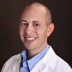 Headshot of a smiling man with medium skin tone and a shaved head, wearing a blue, open-collar dress shirt under a white lab coat against a dark indoor background