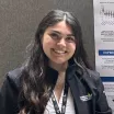 Portrait of a smiling woman with long, wavy black hair and a light skin tone wearing a black CHLA jacket, a black CHLA lanyard, and a light colored blouse standing in front of a research poster