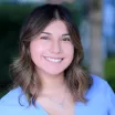 Headshot of a smiling young woman with light skin tone and wavy, shoulder-length brown hair wearing a small silver cross necklace and blue blouse against a blurred outdoor background