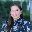 Headshot of a smiling woman with a light skin tone and long brown hair wearing a black blouse with blue floral print against a blurred outdoor background