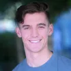 Headshot of a smiling young man with light skin tone and brown short back and sides haircut wearing a blue t-shirt against a blurred outdoor background