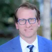 Headshot of a smiling man with light skin tone and short brown hair wearing eyeglasses, a light colored dress shirt and blue tie under a blue suit jacket against a blurred outdoor background