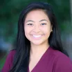 Headshot of a smiling woman with a medium skin tone wearing a maroon scrub top against a blurred outdoor background