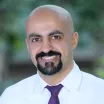 Headshot of a smiling man with medium skin tone, shaved head and dark goatee wearing light dress shirt and purple tie against a blurred outdoor background