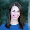 Headshot of a smiling woman with light skin tone and shoulder-length brown hair wearing a solid blue top against a blurred outdoor background