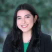 Headshot of a smiling woman with a light skin tone and long black hair wearing a black blazer and green shirt against a blurred outdoor background