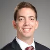 Headshot of a smiling man with light skin tone and short brown hair wearing a white dress shirt and red striped tie under a dark suit jacket against a neutral indoor background