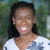 Headshot of a smiling woman with dark skin tone and long dark hair pulled back in braids wearing bright round earrings and a blue floral patterned blouse against a blurred outdoor background