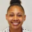 Headshot of a smiling woman with dark skin tone and dark hair pulled back in a bun wearing a white polka dot shirt against a neutral background