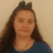 Headshot of a woman with medium skin tone and dark hair tied back with a bow wearing a chain necklace and blue top against a neutral indoor background