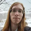 Headshot of a man with light skin tone and lone auburn hair wearing glasses against a snowy outdoor background