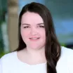 Headshot of a smiling woman with light skin tone and long brown hair wearing white top against a blurred outdoor background