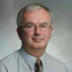 Headshot of a man with light skin tone and short grey hair wearing eyeglasses, grey dress shirt and brown tie against a neutral indoor background