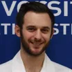 Headshot of a smiling man with light skin tone and dark hair and beard wearing a white lab coat against a blue background with indistinguishable white lettering