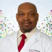 Headshot of a man with dark skin tone, shaved head and dark goatee wearing a white dress shirt and red tie under a white lab coat against a light patterned indoor background