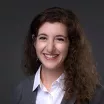 Headshot of a smiling woman with light skin tone and wavy brown hair wearing a light colored blouse under a dark suit jacket against a dark indoor background