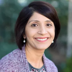 Headshot of a woman with medium skin tone and dark hair wearing a purple sweater against a blurred outdoor background