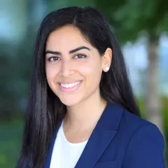 Headshot of young woman with medium skin tone and dark hair wearing business attire against blurred outdoor background